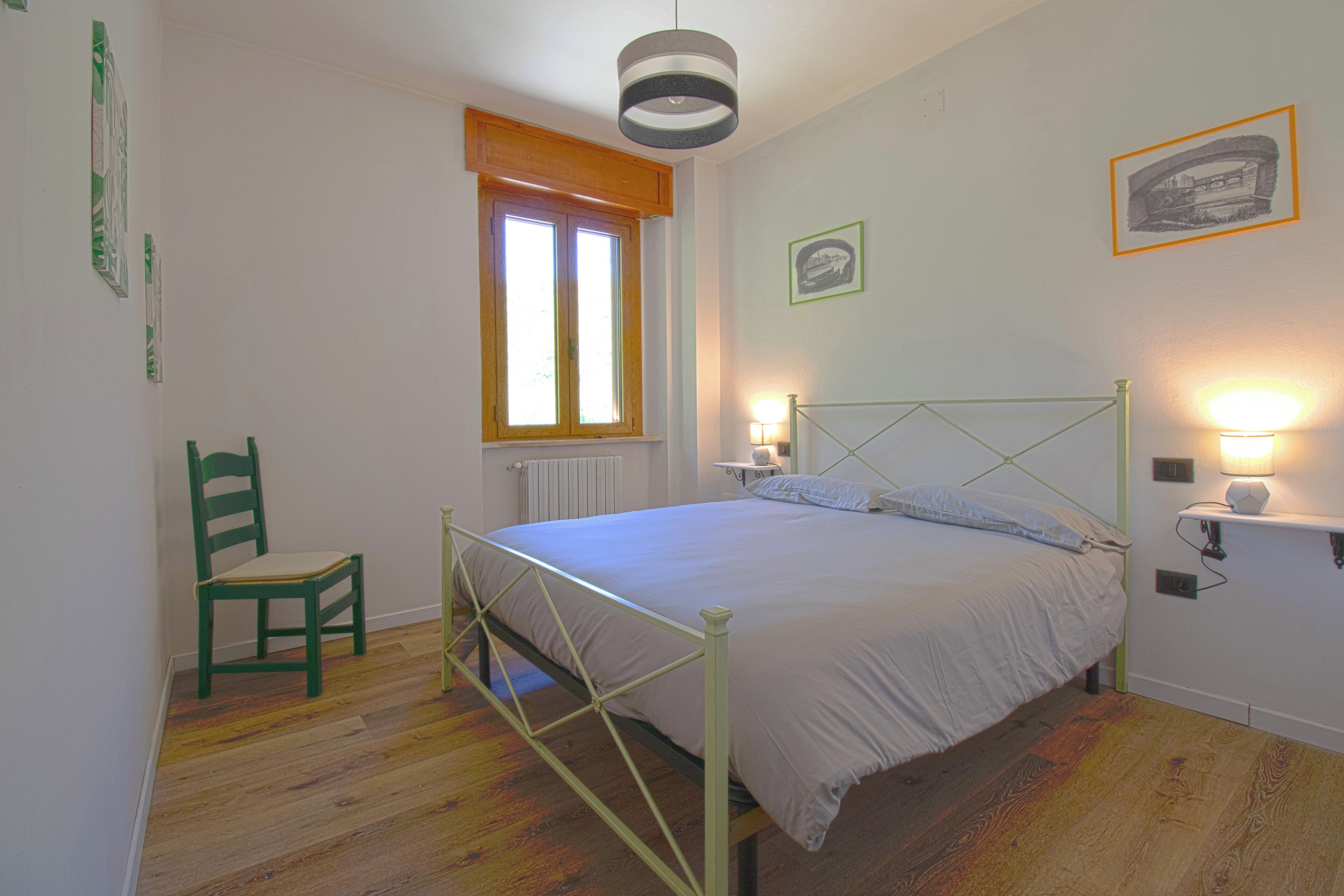 Un dormitorio con cama, una silla, un ventanal y luces fijadas a la pared.