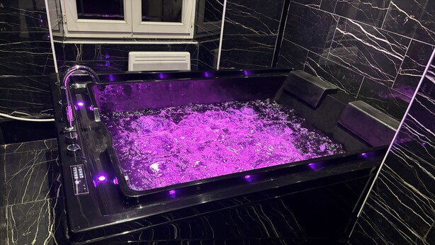 A black marble bathtub with purple lighting, a window above, and a white vent.