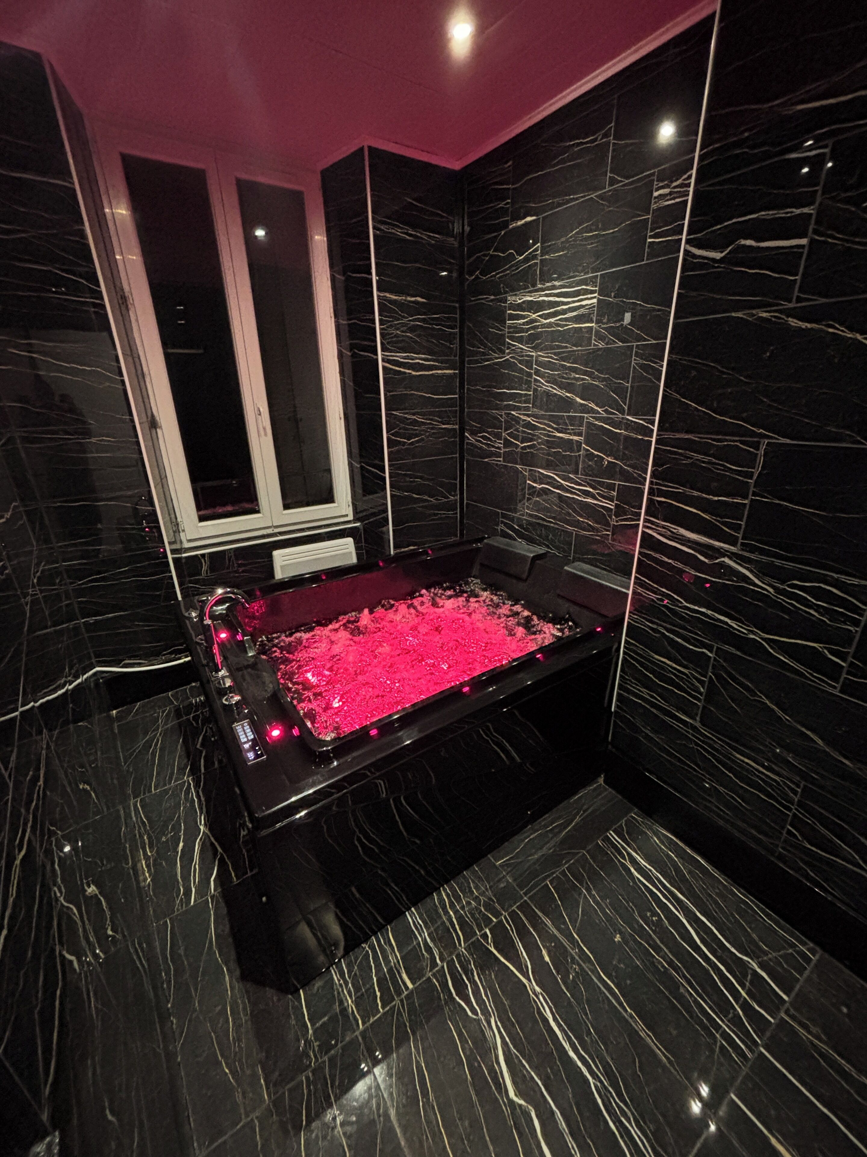 A modern bathroom with a black marble bathtub, a window, and a pink-lit floor.