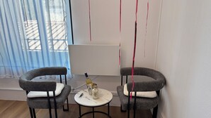 A room with two grey upholstered chairs, a small round table, a bottle of wine, and a salt shaker.