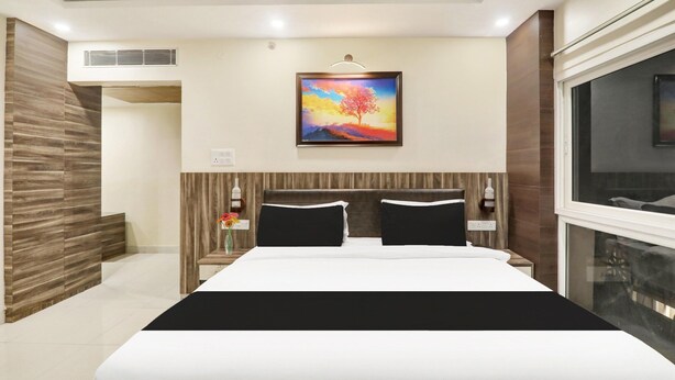 A modern hotel room with a large bed, a painting above the bed, a nightstand, a window, and a wooden accent wall. - Deluxe Kunwar Aalcajars (Patna)