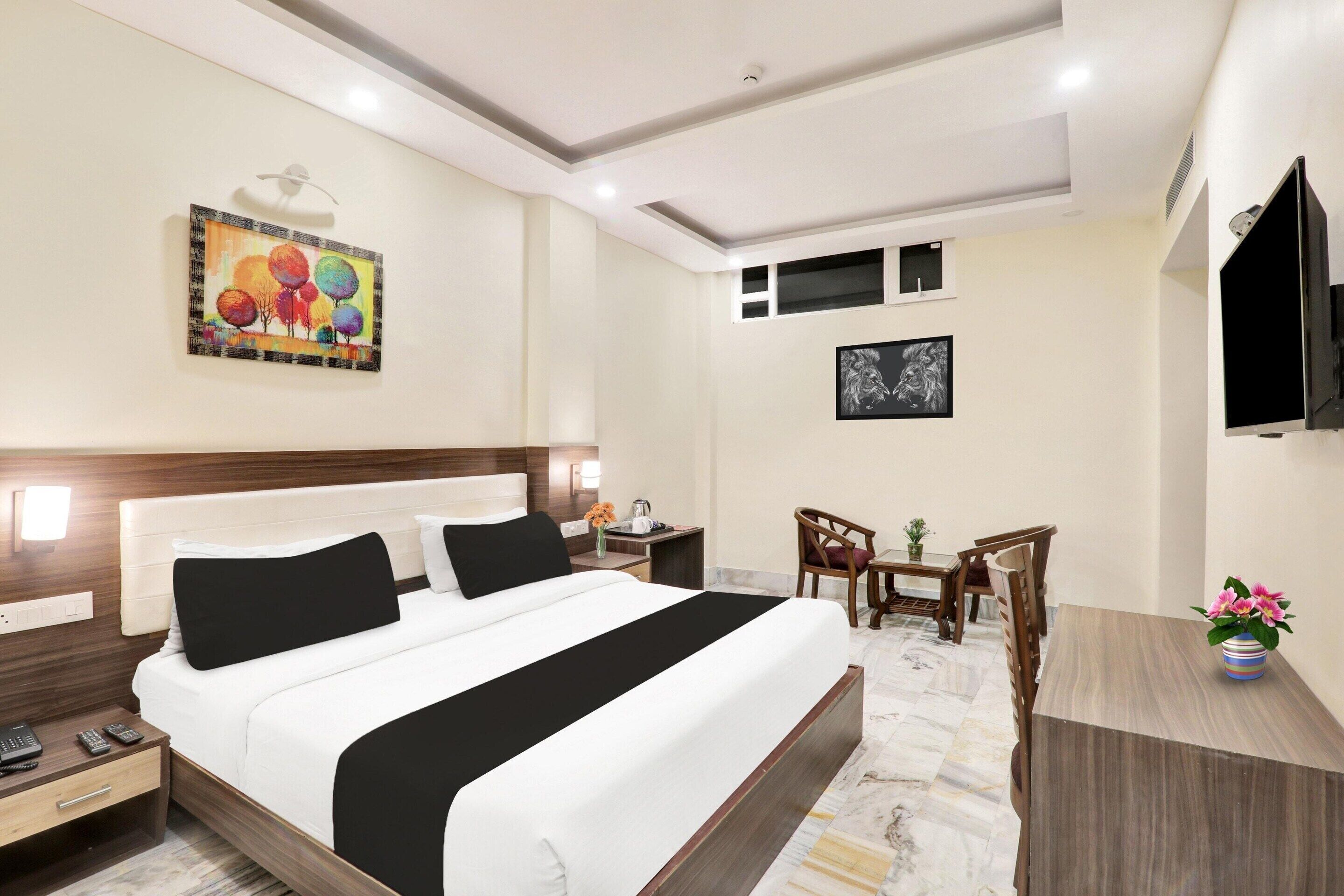 A hotel room with a bed, bedside tables, a desk with chairs, a television, and artwork on the walls.