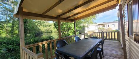 A covered outdoor deck with a table and chairs, surrounded by lush greenery and a view of a house.