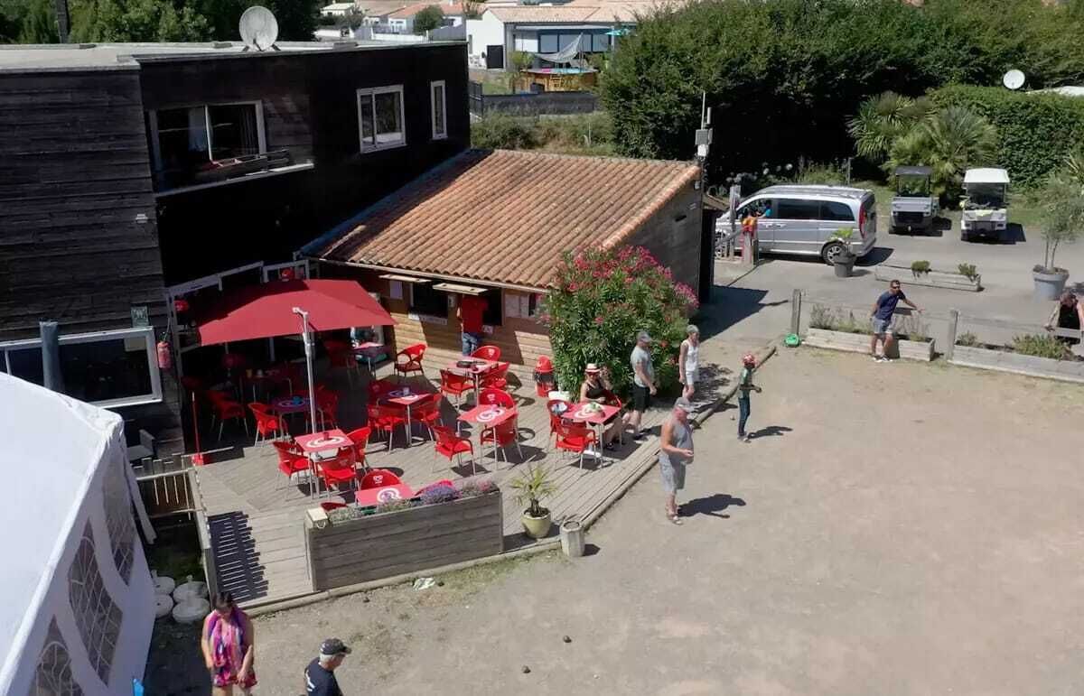 Ein Sitzbereich im Freien mit roten Tischen und Stühlen, ein Gebäude mit einem Ziegeldach und ein Parkplatz mit Fahrzeugen.