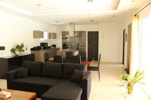 Un salon moderne avec un canapé d’angle, un coin repas doté d’une table en verre et une cuisine équipée d’appareils électroménagers intégrés.