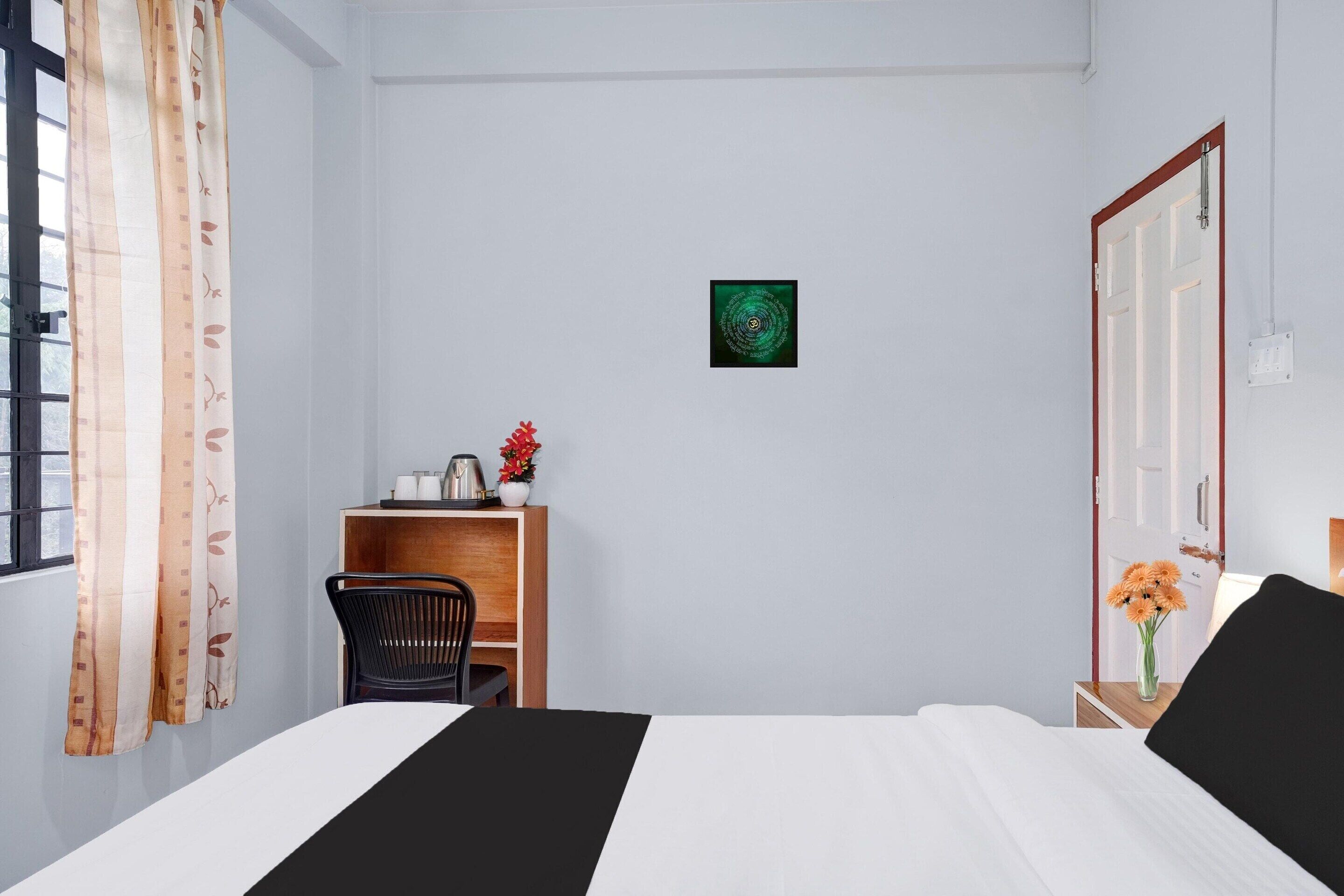 A hotel room with a bed, a desk with a chair, a small table with a vase of flowers, and a wall-mounted picture.