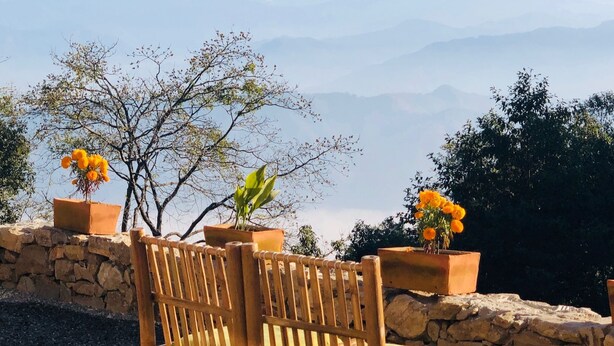 Un banco de madera frente a un muro de piedra y macetas con flores, con vistas a una cordillera.