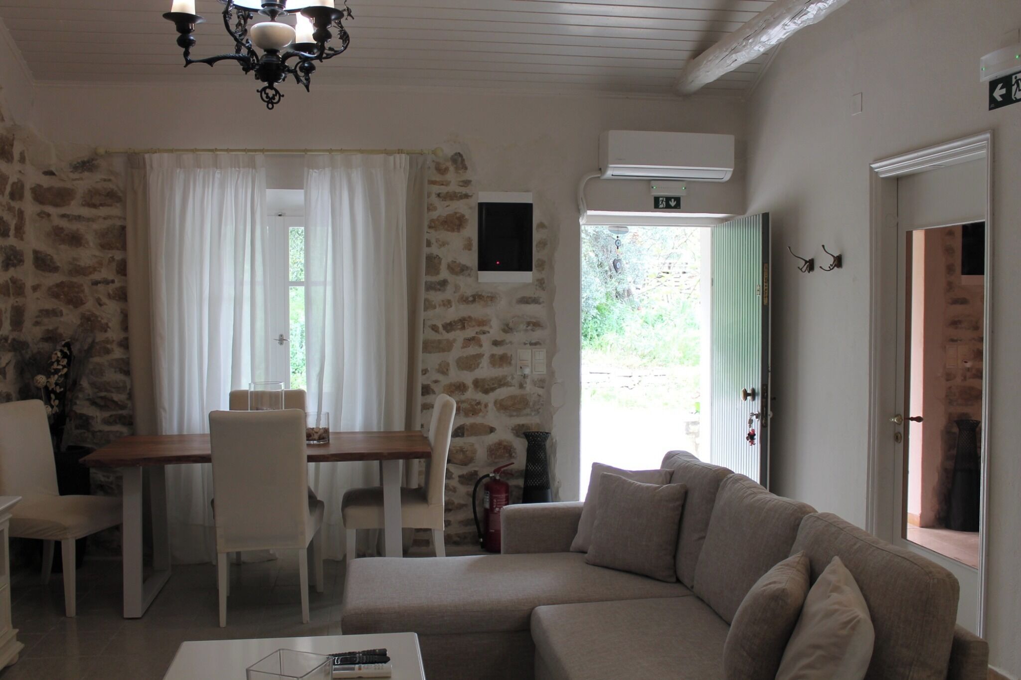 Una stanza con parete in pietra, un tavolo di legno con sedie, un divano color beige, un lampadario e un estintore.