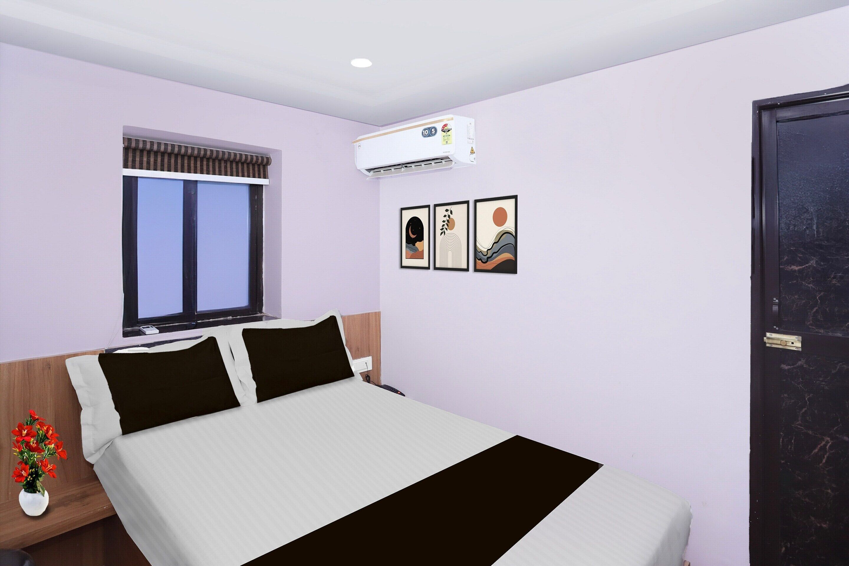 A hotel room with a bed, a window with blinds, an air conditioning unit, and three framed pictures on the wall.