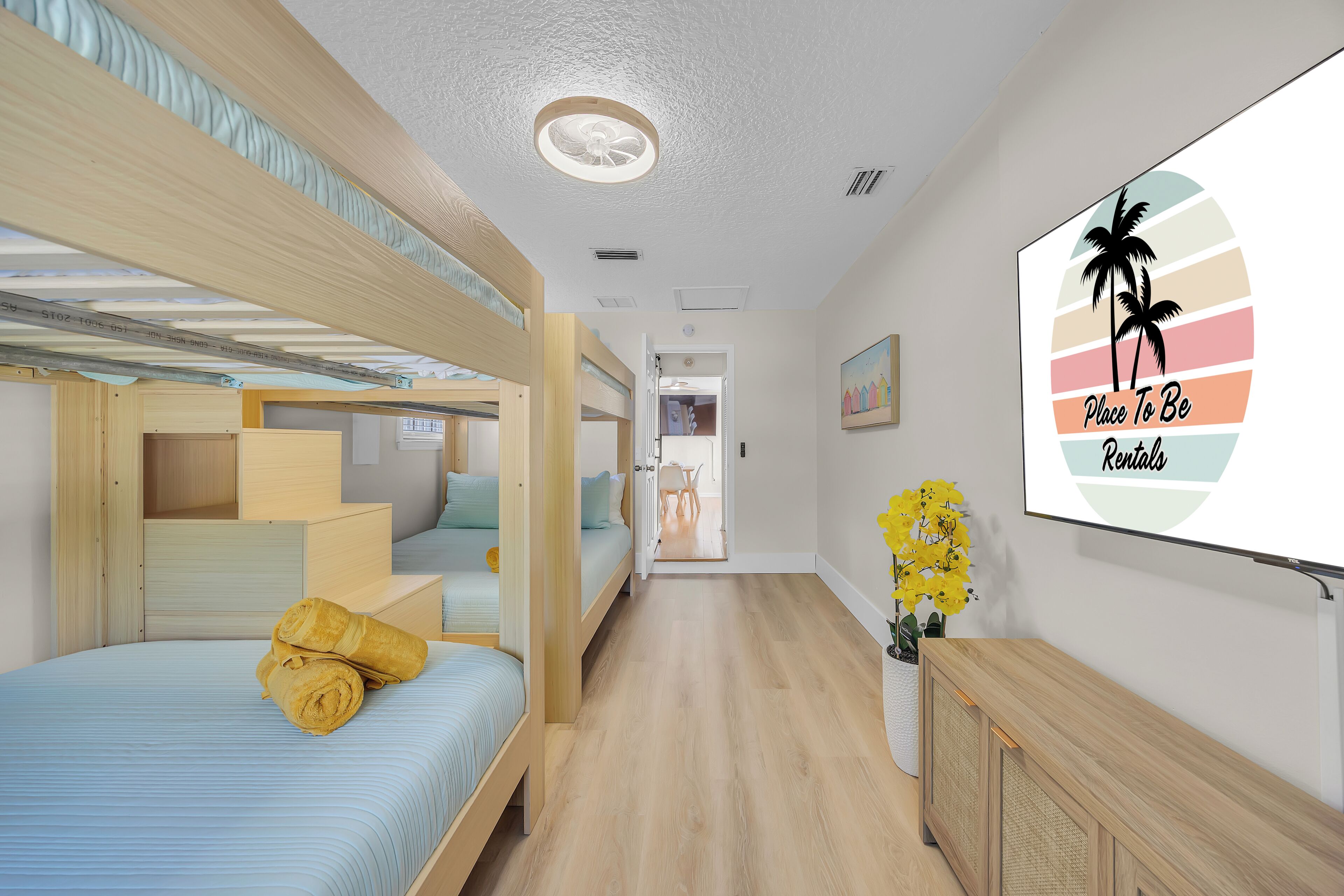 A room with bunk beds, a wooden cabinet, a wall-mounted TV, and a hallway leading to another room.