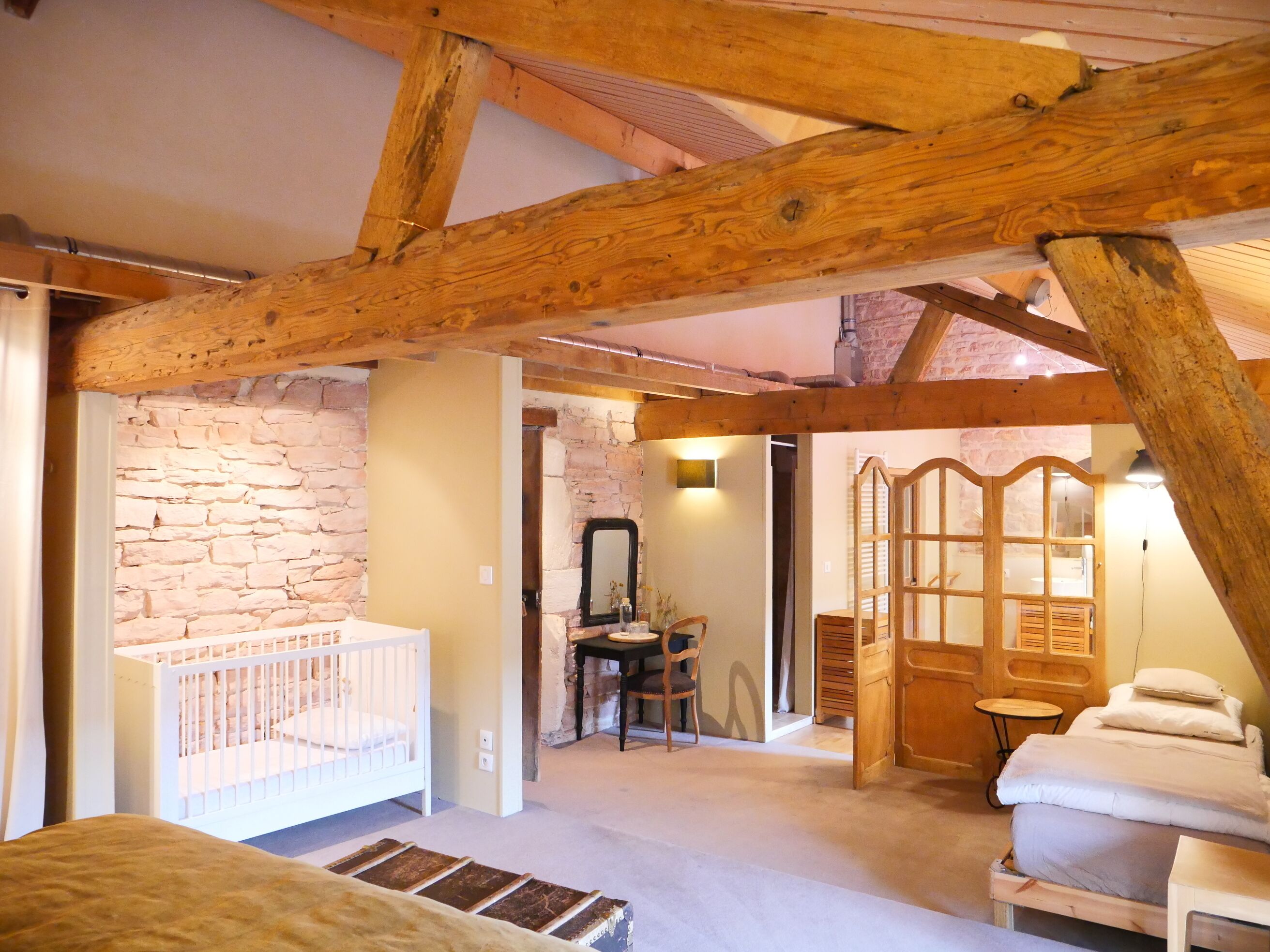 Une pièce rustique avec des poutres apparentes, un mur en pierre, un berceau, une porte en bois et un lit.