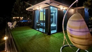 Una estructura moderna con paredes de vidrio y techo plano, un sillón colgante y césped artificial verde.