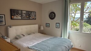 Ett sovrum med en säng, ett nattduksbord, en lampa, ett fönster med utsikt över grönområden och konst på väggen.