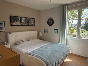 A bedroom with a bed, a nightstand, a lamp, a window with a view of greenery, and wall art. - Aix en Provence, near, beautiful country house 200 m2 with independent studio and swimming pool (VENTABREN)