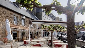 Un café al aire libre con mesas y sillas, un edificio de piedra con un letrero, y un árbol que da sombra.