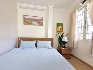 Un dormitorio con una cama de madera, una mesita de noche con lámpara, un cuadro y una ventana con cortinas.