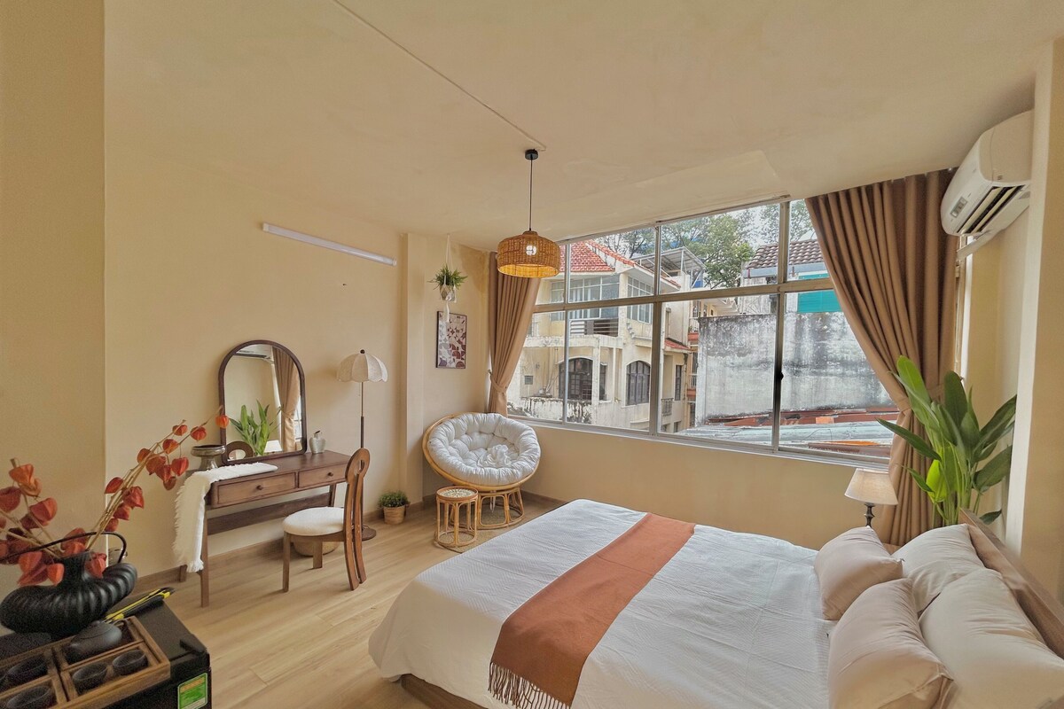 A bedroom with a bed, a desk, a chair, a hanging light fixture, a window with curtains, and an air conditioning unit.