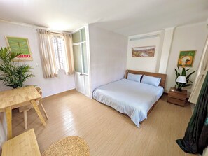Un dormitorio con una cama, una mesa de madera, una silla, un ventanal con cortinas y un cuadro en la pared.