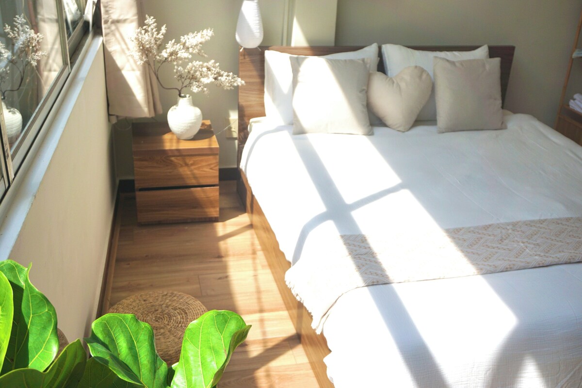 A bedroom with a wooden bed, white bedding, a bedside table with a vase, and a window with natural light.