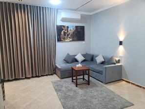 Una sala de estar con un sofá gris, una pequeña mesa de madera, un tapete gris, un cuadro en la pared y una ventana con cortinas.