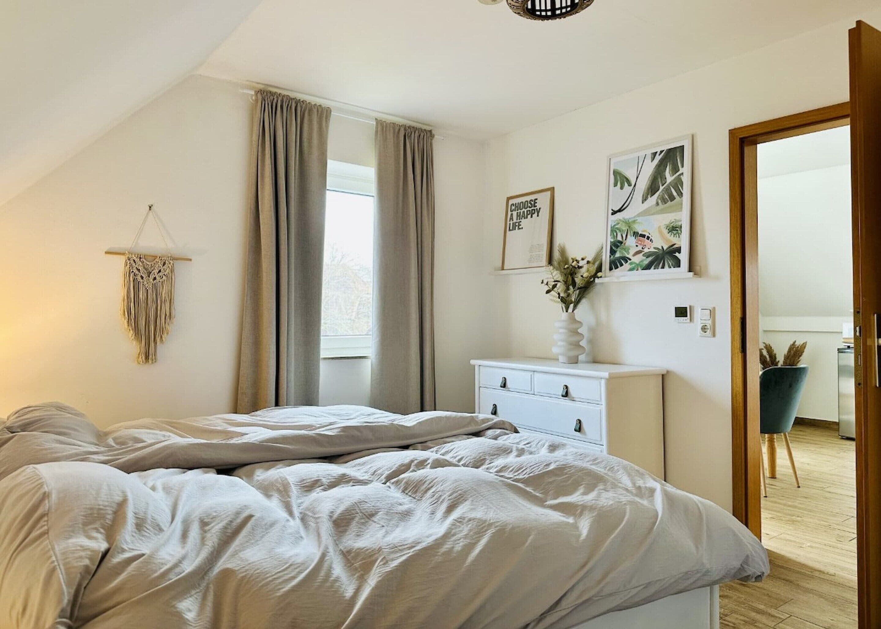 A bedroom with a bed, a dresser, a macrame wall hanging, and a window with curtains.