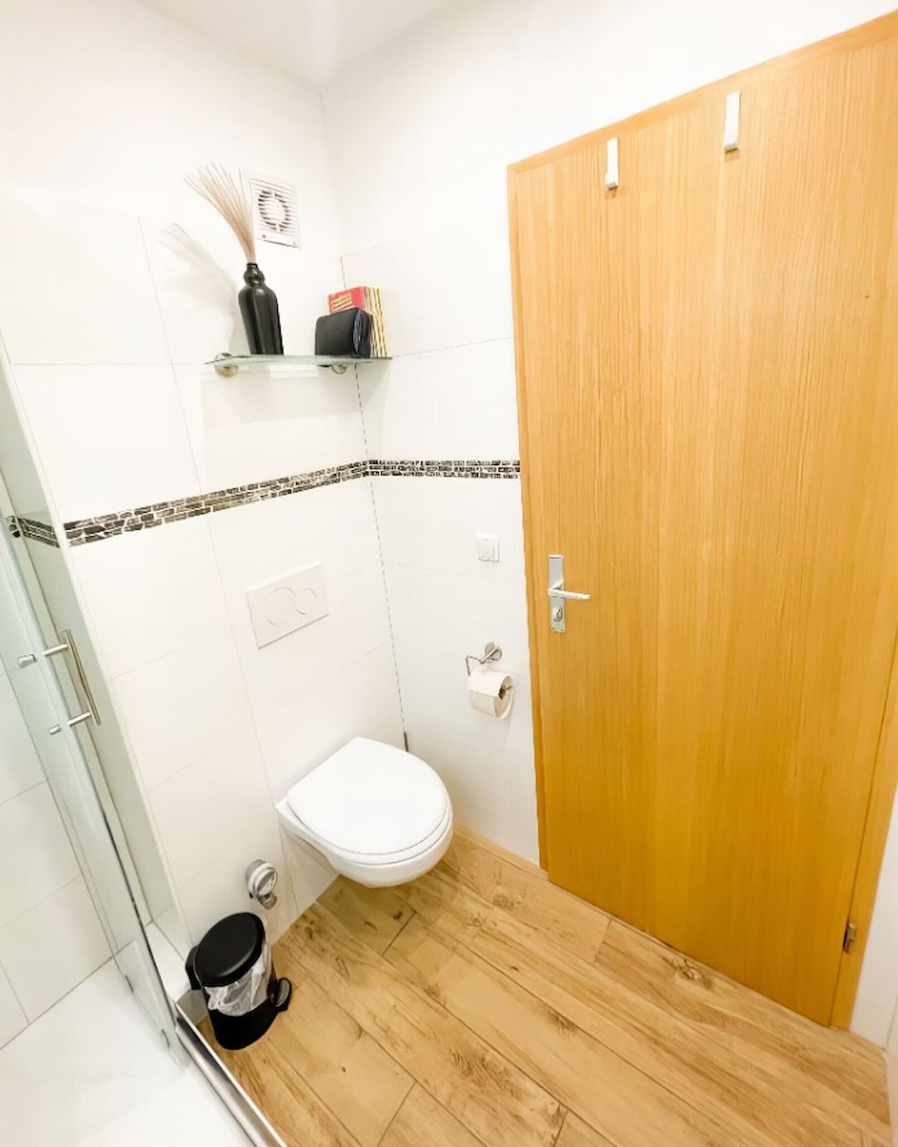Un baño con inodoro, ducha, puerta de madera y estante con artículos de aseo.