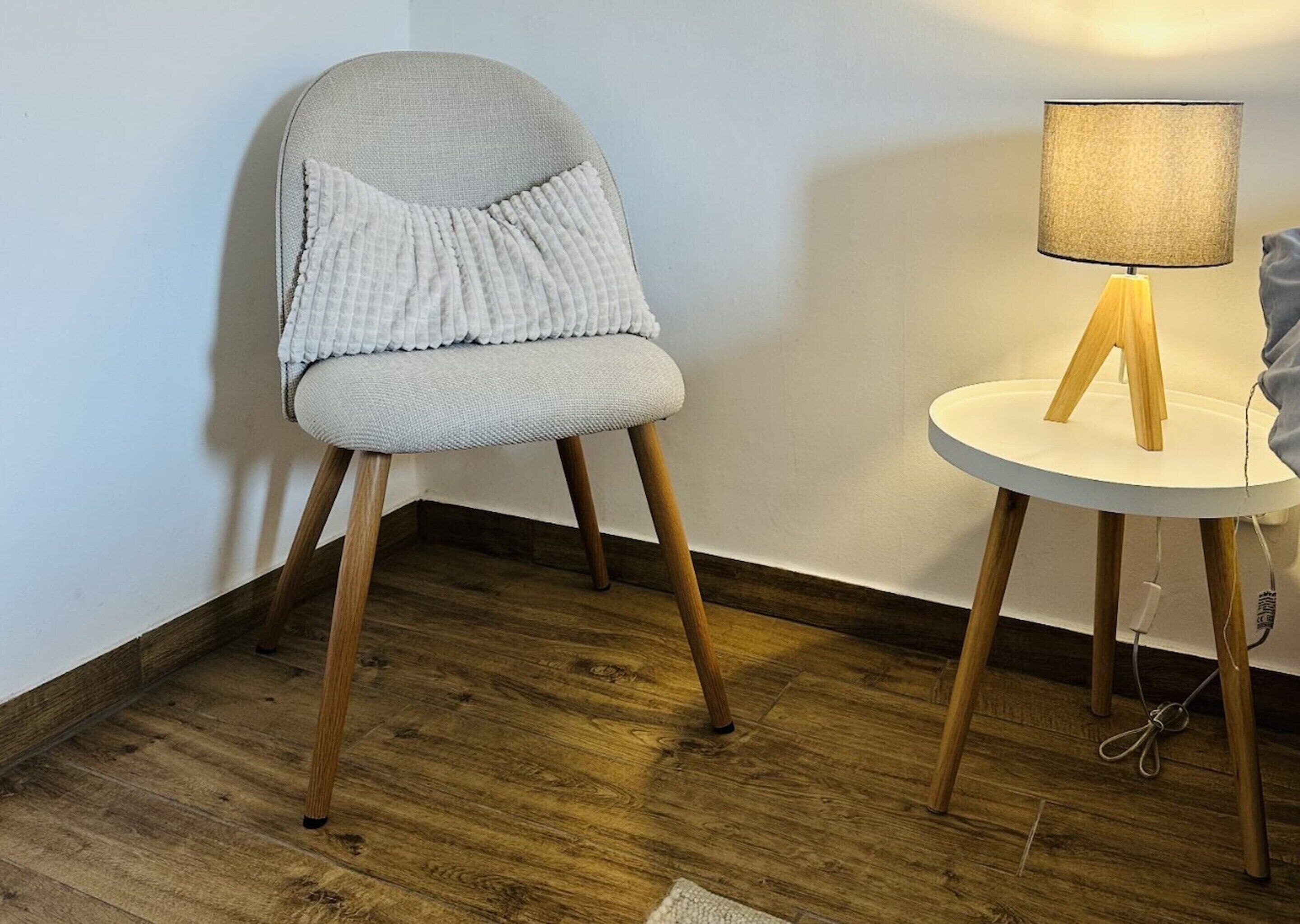 A chair with a textured seat and wooden legs, a small round table with a lamp, and a wooden floor.