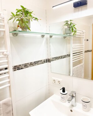 Un baño con repisa de vidrio que sostiene una planta, un espejo, un lavabo con grifo y un perchero para toallas.