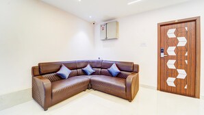 A corner living room with a brown leather sofa, a wooden door with geometric patterns, and a ceiling-mounted air conditioning unit. - Deluxe Room Super Hotel O Aarapalayam (Madurai)