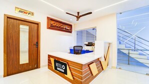 A hotel reception desk with a wooden finish, a ceiling fan, a framed artwork, and a staircase in the background. - Deluxe Room Super Hotel O Aarapalayam (Madurai)