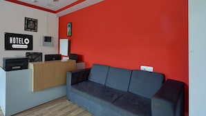 A hotel reception area with a reception desk, a grey sofa, a ceiling fan, and a red wall. - Deluxeurban Drift (Bengaluru)