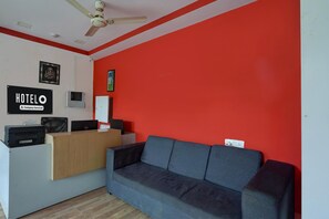 Ein Hotel-Empfangsbereich mit einer Rezeption, einem grauen Sofa, einem Deckenventilator und einer roten Wand.
