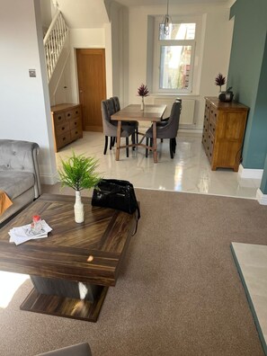 Ein modernes Wohnzimmer mit einem Esstisch, Stühlen, einer Couch, einem Couchtisch mit Vase und Handtasche sowie einer Treppe.