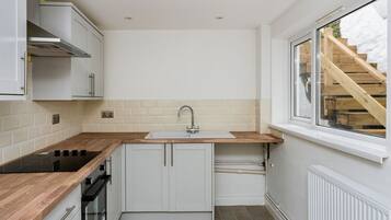 A compact kitchen with a wooden countertop, white cabinets, a built-in stove, and a window with a view of a wooden staircase.