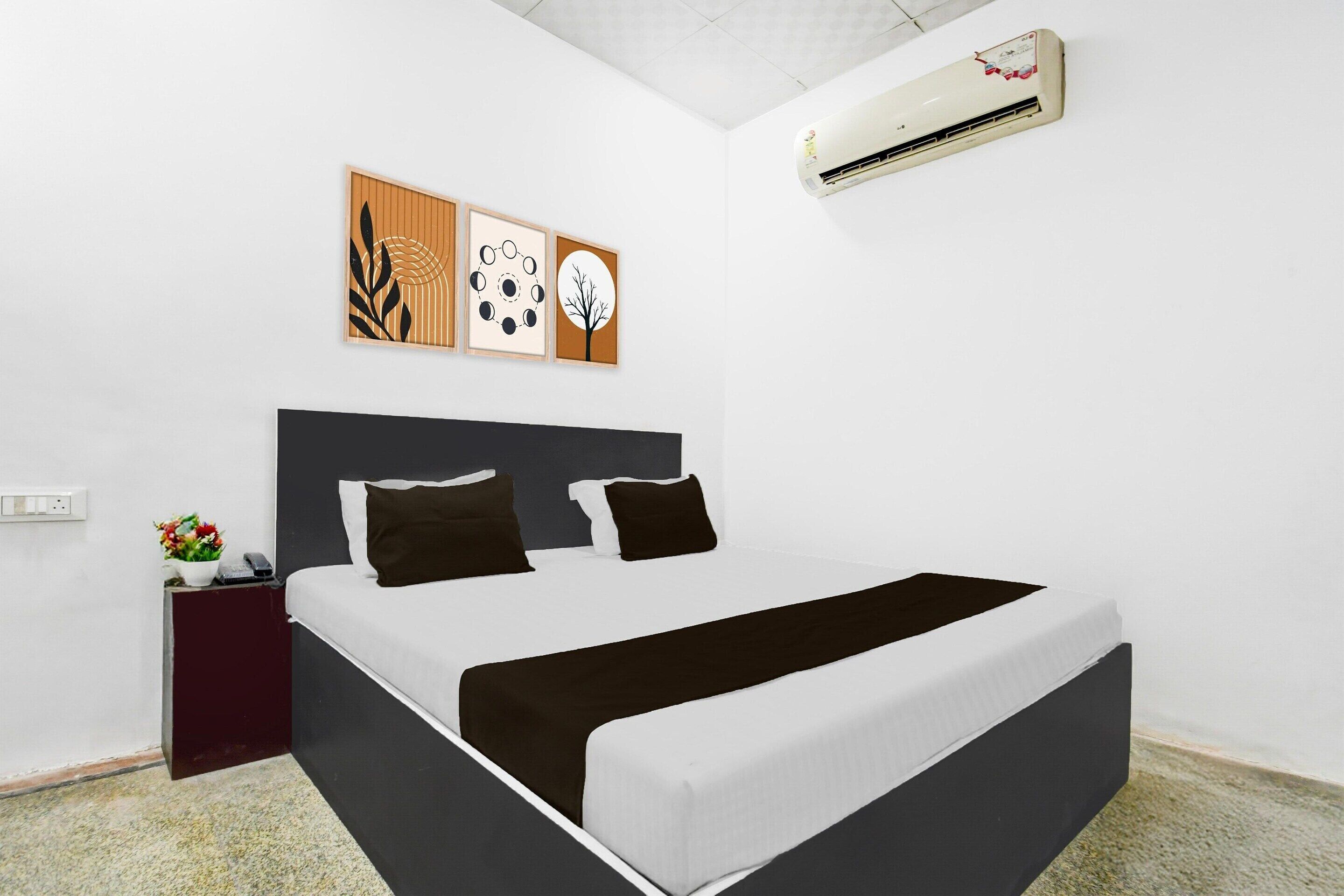 A modern hotel room with a large bed, a dark headboard, a wall-mounted air conditioner, and a small side table with a vase of flowers.