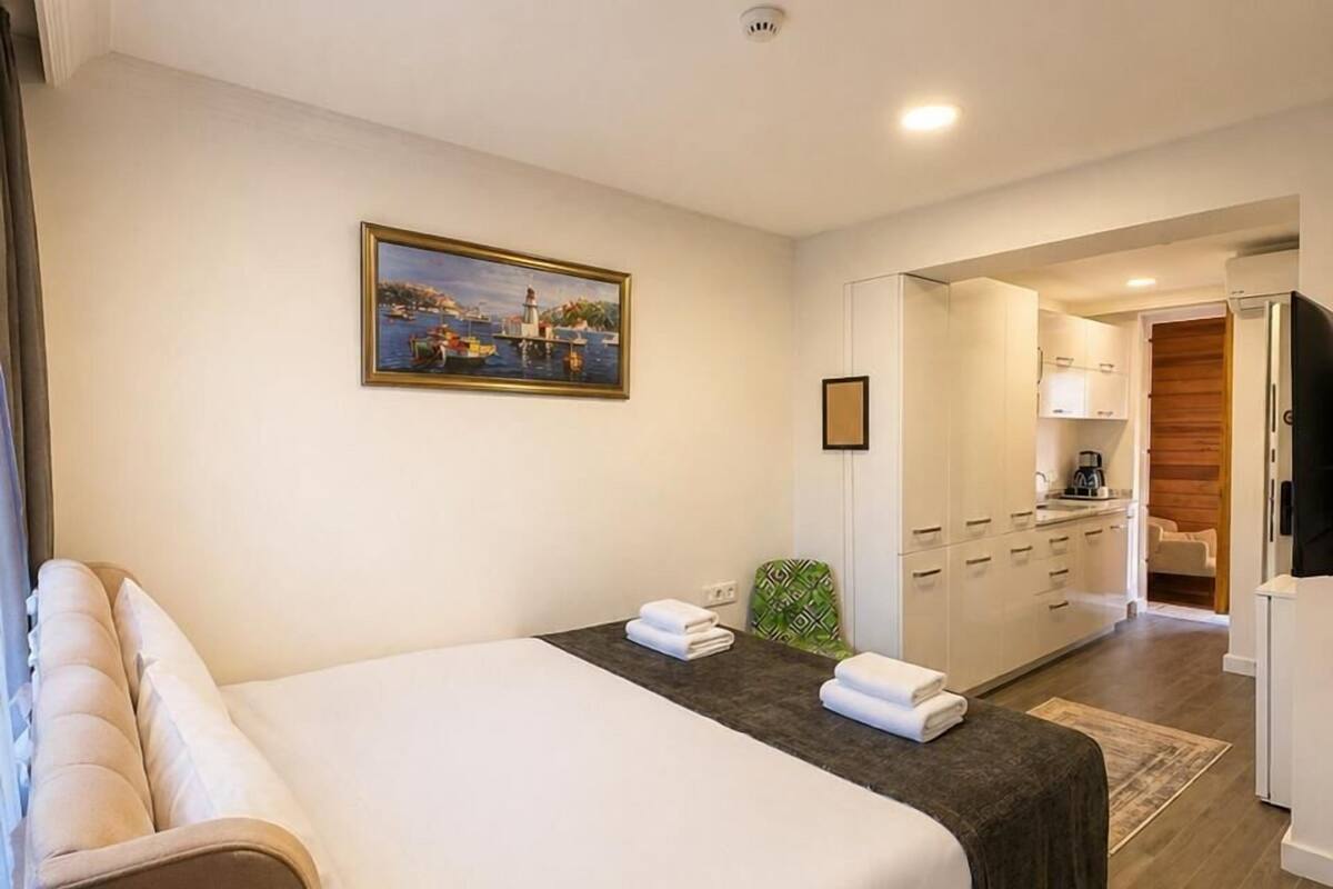 A hotel room with a bed, a small table with a remote control, a painting on the wall, and a view into the kitchen area.