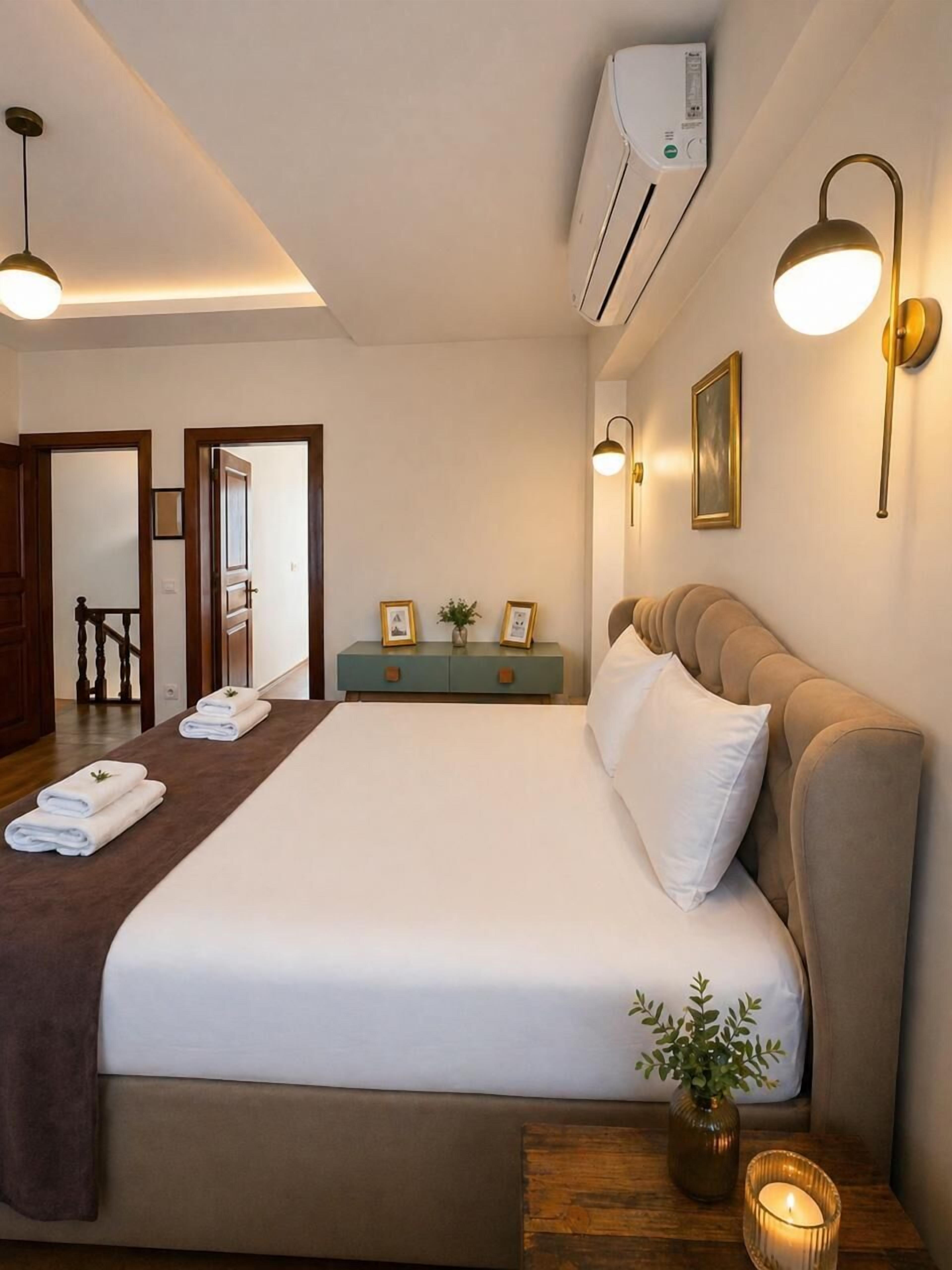 A hotel room with a bed, bedside lamps, a small table with a candle, a vase with a plant, and an air conditioning unit.