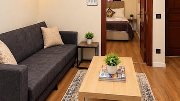 Een moderne woonkamer met een donkergrijze bank, een houten salontafel met een plant en tijdschriften, een klein bijzettafeltje, en een gang die leidt naar een slaapkamer met een bed en nachtkastje.