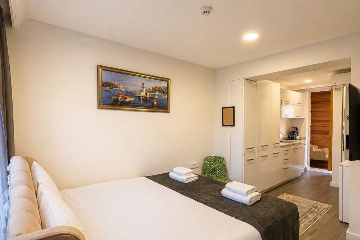 A hotel room with a bed, a small table with a remote control, a painting on the wall, and a kitchenette in the background.