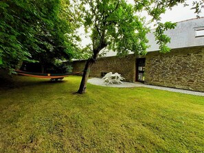 Une cour verdoyante avec un hamac, un arbre et un bâtiment en pierre doté d’une porte et d’une fenêtre.