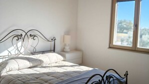 A bedroom with a wooden ceiling, a large window, a bed with a metal frame, and a bedside table with a lamp. - A dwelling where time slows down and regain the rhythm of nature. (Morrovalle)