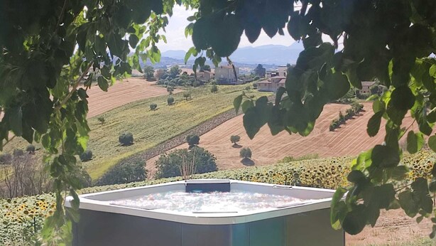 A hot tub in an outdoor setting with a view of rolling hills and a distant village. - A dwelling where time slows down and regain the rhythm of nature. (Morrovalle)