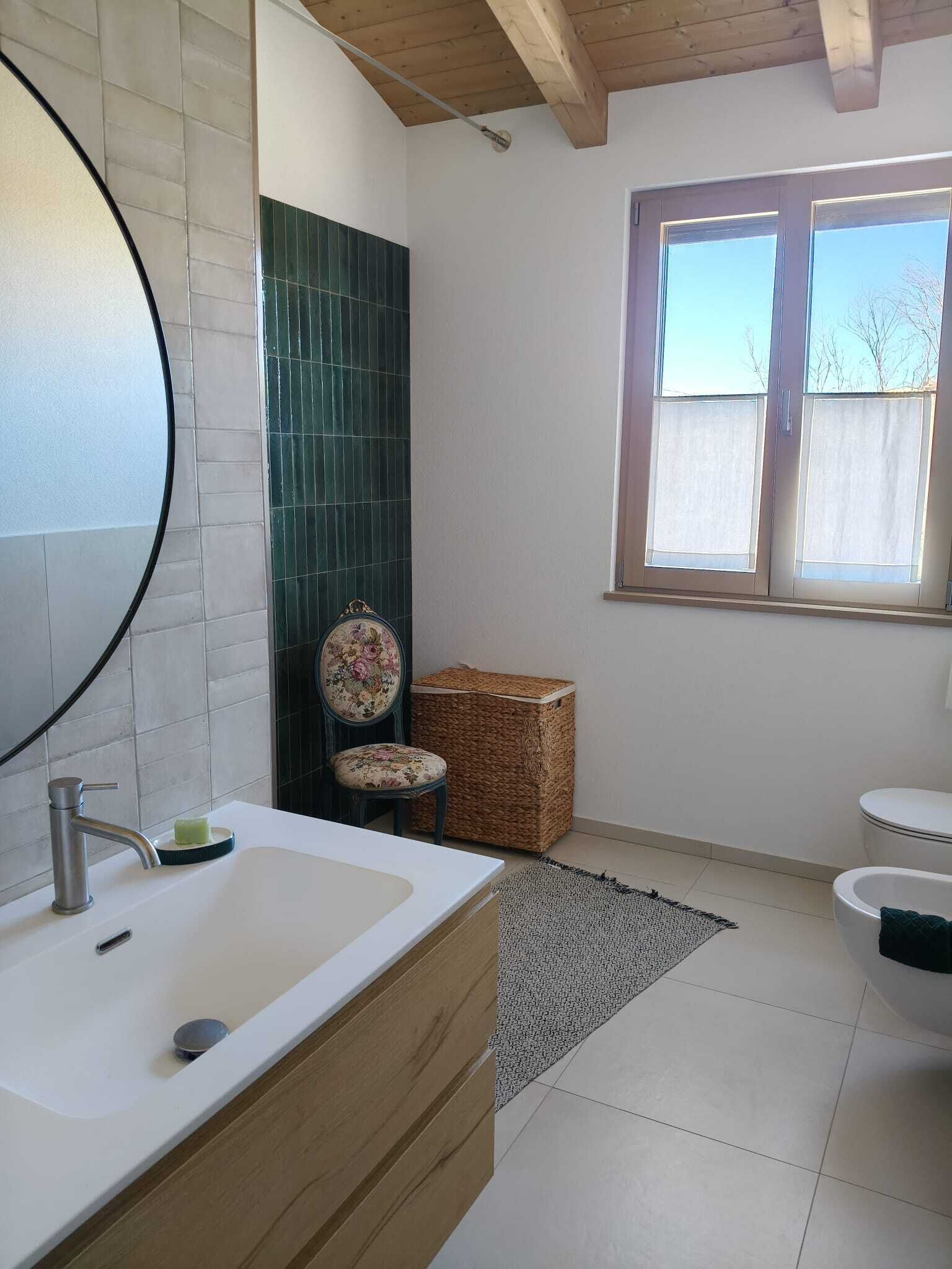 Ein Badezimmer mit einem großen Spiegel, einem Holzkosmetikschrank, einer grüne gefliesten Dusche, einem Korbwannen und einem Fenster mit Blick.