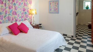 Un dormitorio con piso a cuadros, una cama con ropa blanca y dos almohadas rosas, una mesita de noche con lámpara, un cuadro en la pared y una ventana con una planta.
