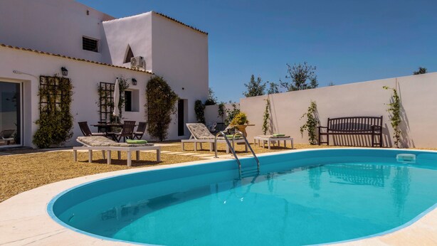 Una piscina con escalera, sillas de descanso y un edificio blanco de techo azulejado.