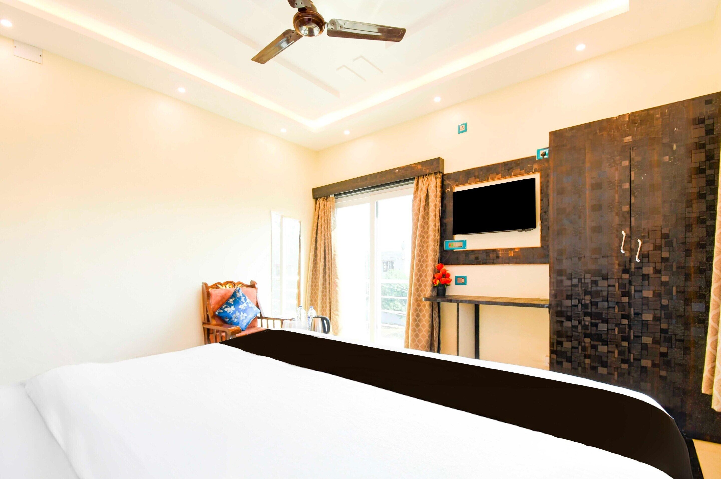 Habitación de hotel con cama, ventilador de techo, televisión, armario y ventana con cortinas.