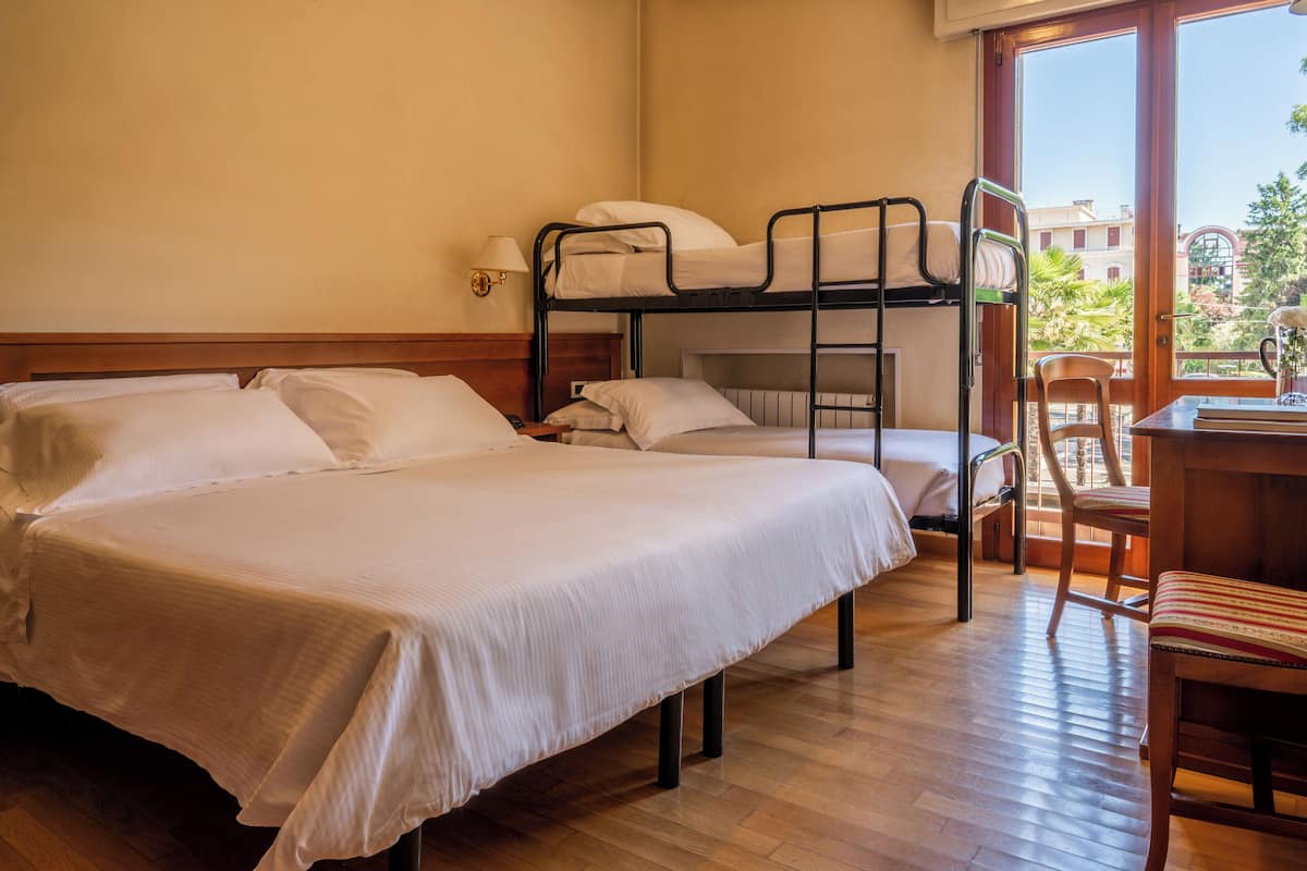 Hotelkamer met een stapelbed, een eenpersoonsbed, een bureau met stoel en een raam met uitzicht op gebouwen en bomen.