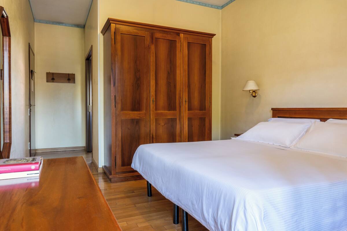 Een slaapkamer met een houten garderobe, een bed met witte lakens en een houten tafel met boeken.