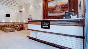 A reception area with a wooden reception desk, a framed cityscape painting, a statue, and a seating area with sofas and a small table. - Deluxe Hotel TR (Amritsar)