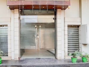 A building entrance with a glass door, a wooden overhang, and two potted plants on the ground.
